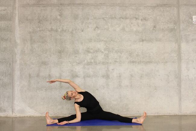Blonde woman with grey background doing side stretch in a split stance on a blue mat at MSMCHQ