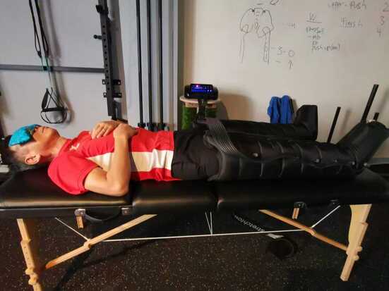 Man in red tee shirt lying down on table with eye pillow getting air compression therapy suit on legs at movement & sports Medicine Centre